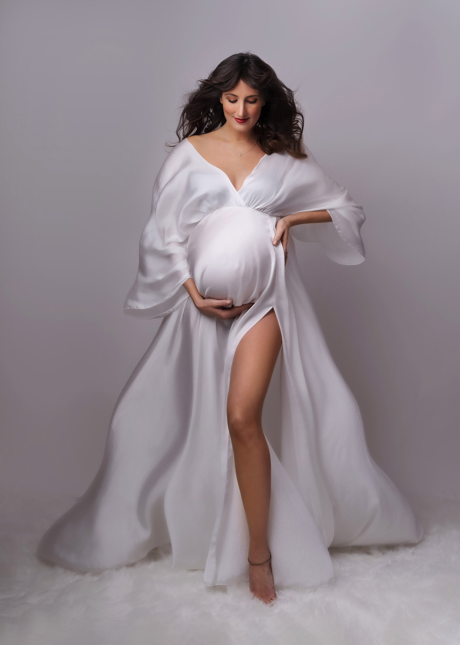 pregnant woman with long dark hair in a white flowing silk dress by maternity photographer in Surrey